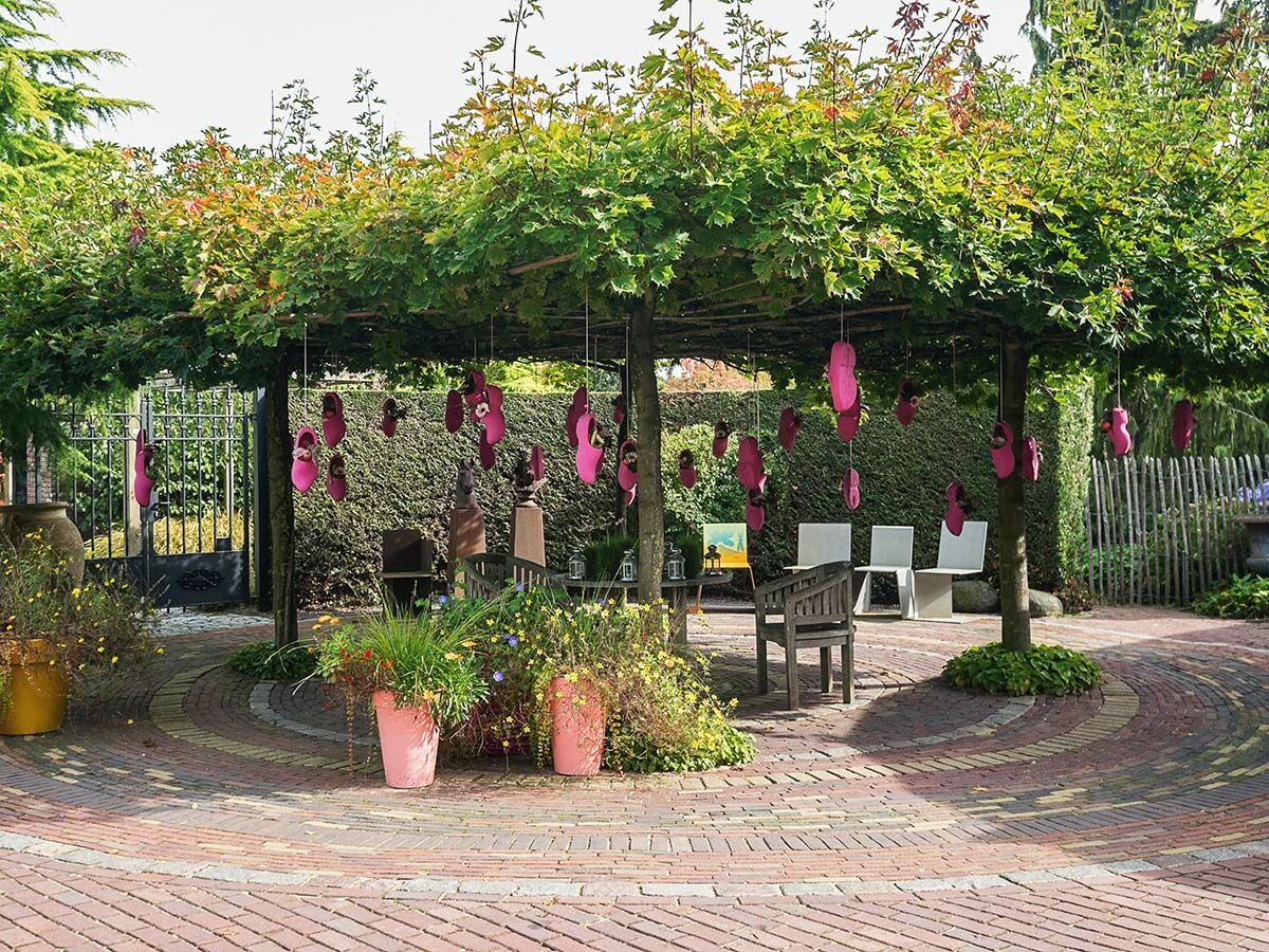 Gartenhof mit baumbestandenem Baldachin, roten Ziegeln und pinkfarbenen Deko-Schuhen, umgeben von Pflanzen und Sitzgelegenheiten.