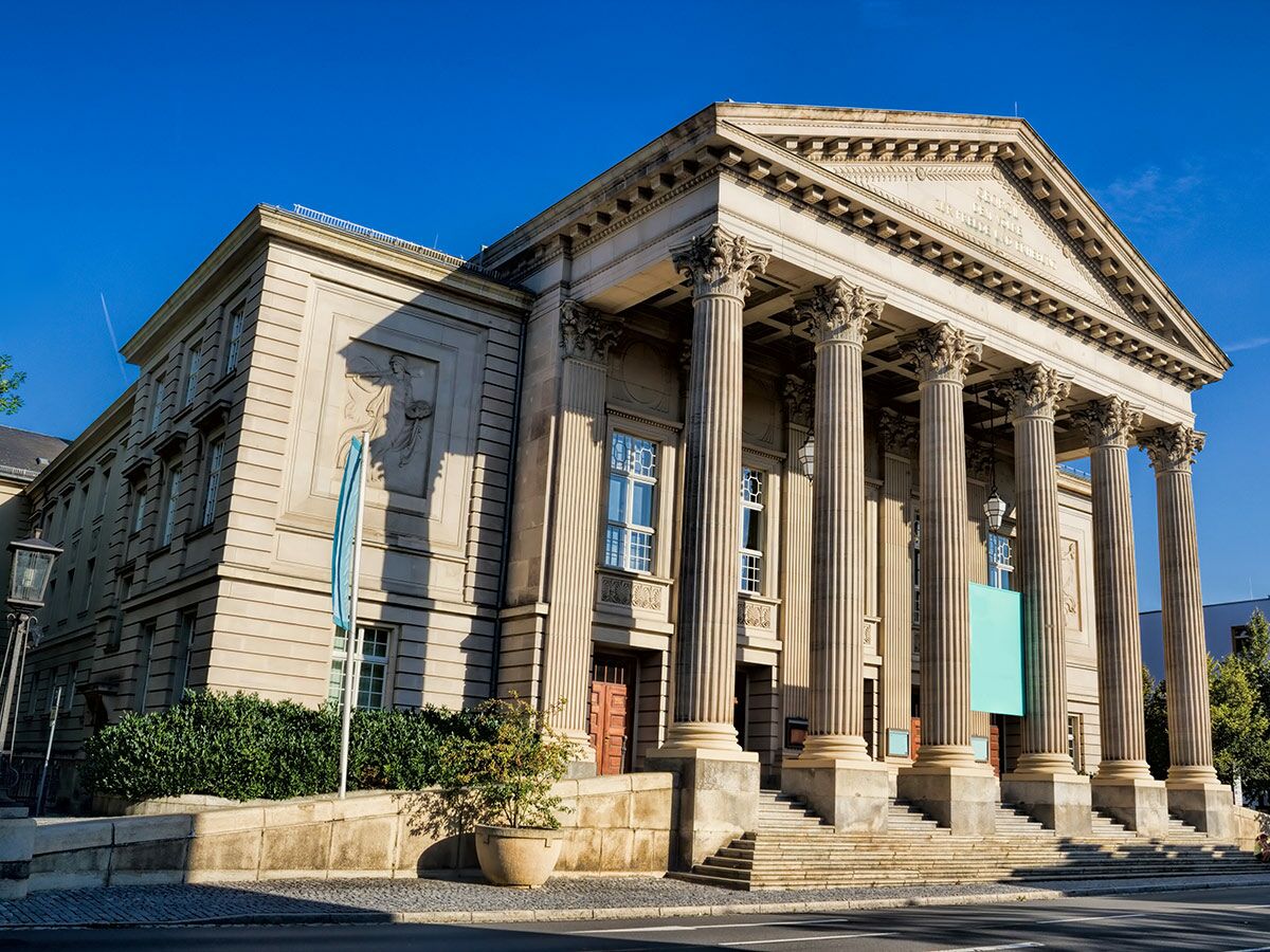 Majestätisches klassisches Gebäude mit großen Säulen, Giebeldreieck und breiter Treppe unter blauem Himmel. Wahrscheinlich Theater oder Museum.