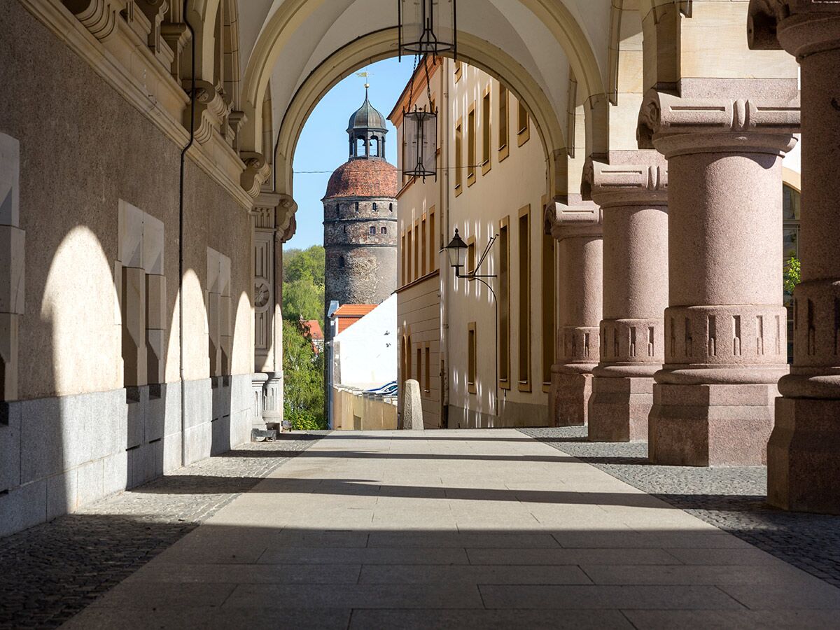 Blick durch einen historischen steinernen Bogen mit Säulen und Schatten auf einen mittelalterlichen Turm mit rotem Dach in einer europäischen Altstadt.