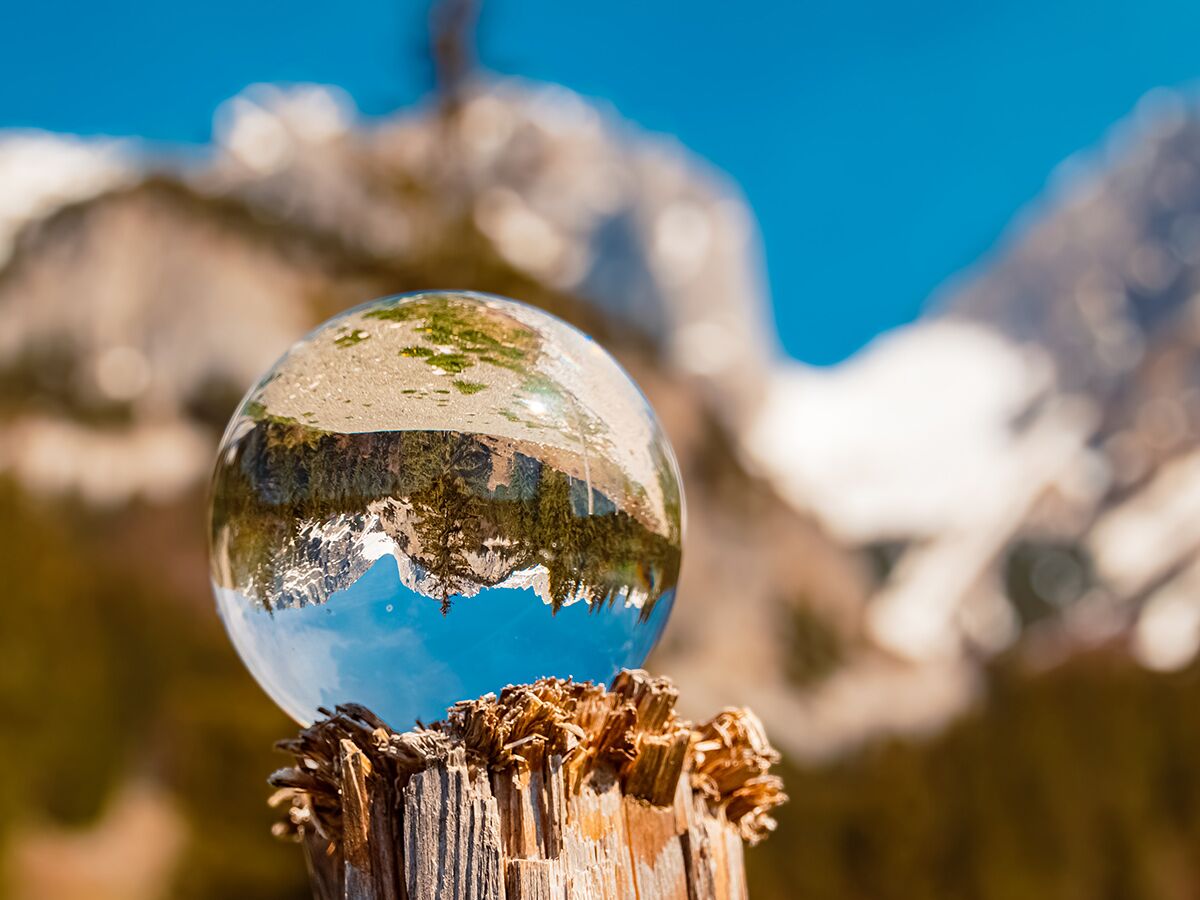 Eine Kristallkugel auf einem Holzpfahl zeigt eine auf dem Kopf stehende Spiegelung einer verschneiten Berglandschaft unter blauem Himmel.