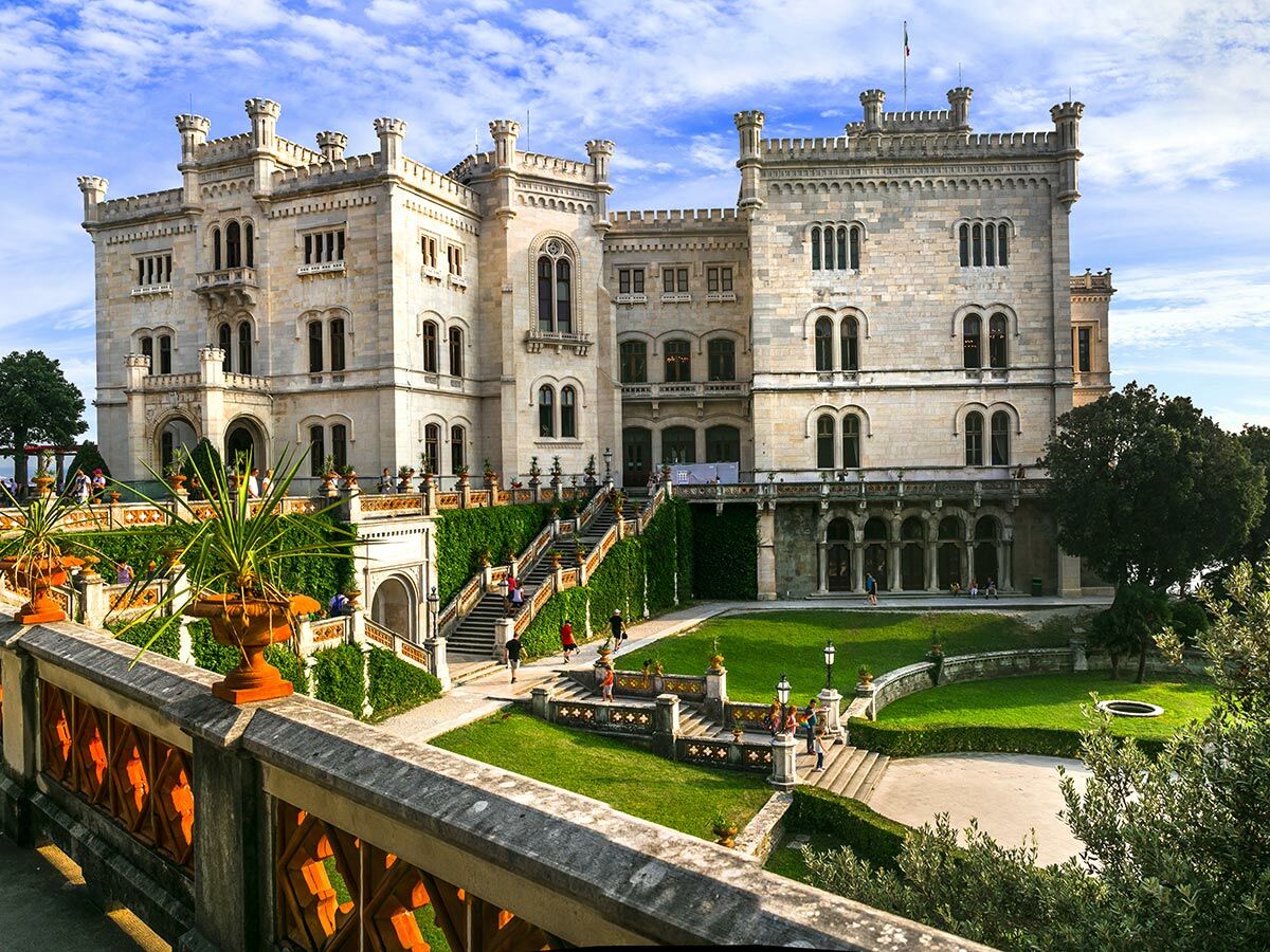 Historisches Schloss Miramare mit Gärten, Treppen und Besuchern unter blauem Himmel in Triest, Italien.