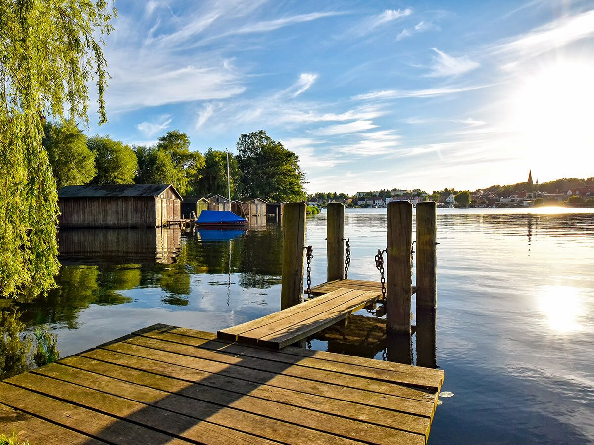 Blick auf einen Holzsteg an einem See mit Bootshäusern und einem Weidenbaum im Abendlicht. Im Hintergrund ist ein Dorf zu sehen.