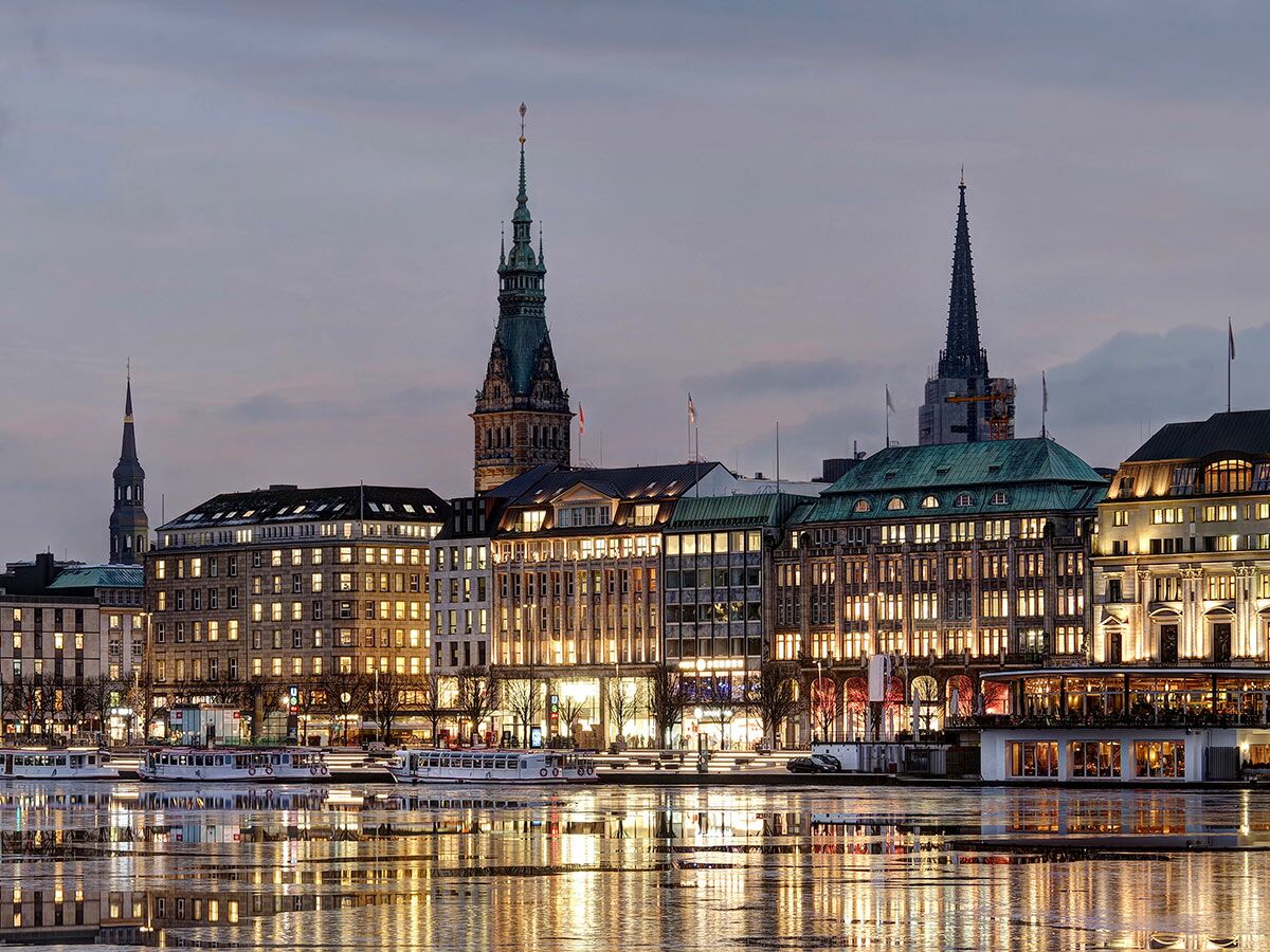 Abendlicher Blick auf die Hamburger Alster mit beleuchteten Gebäuden, spiegelnden Lichtern im Wasser und den Türmen des Rathauses.