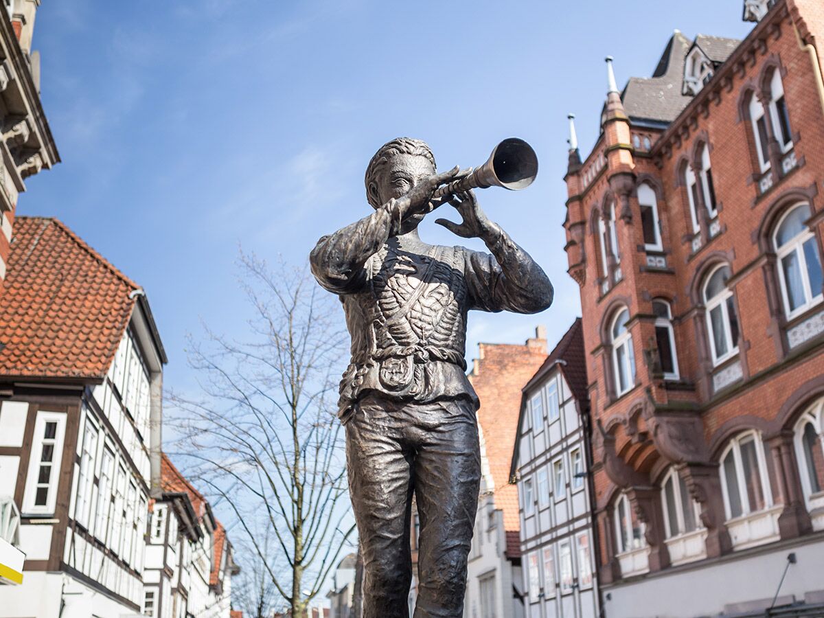 Bronzestatue eines Hornisten vor historischen Fachwerkhäusern und einem roten Backsteingebäude in einer europäischen Altstadt.