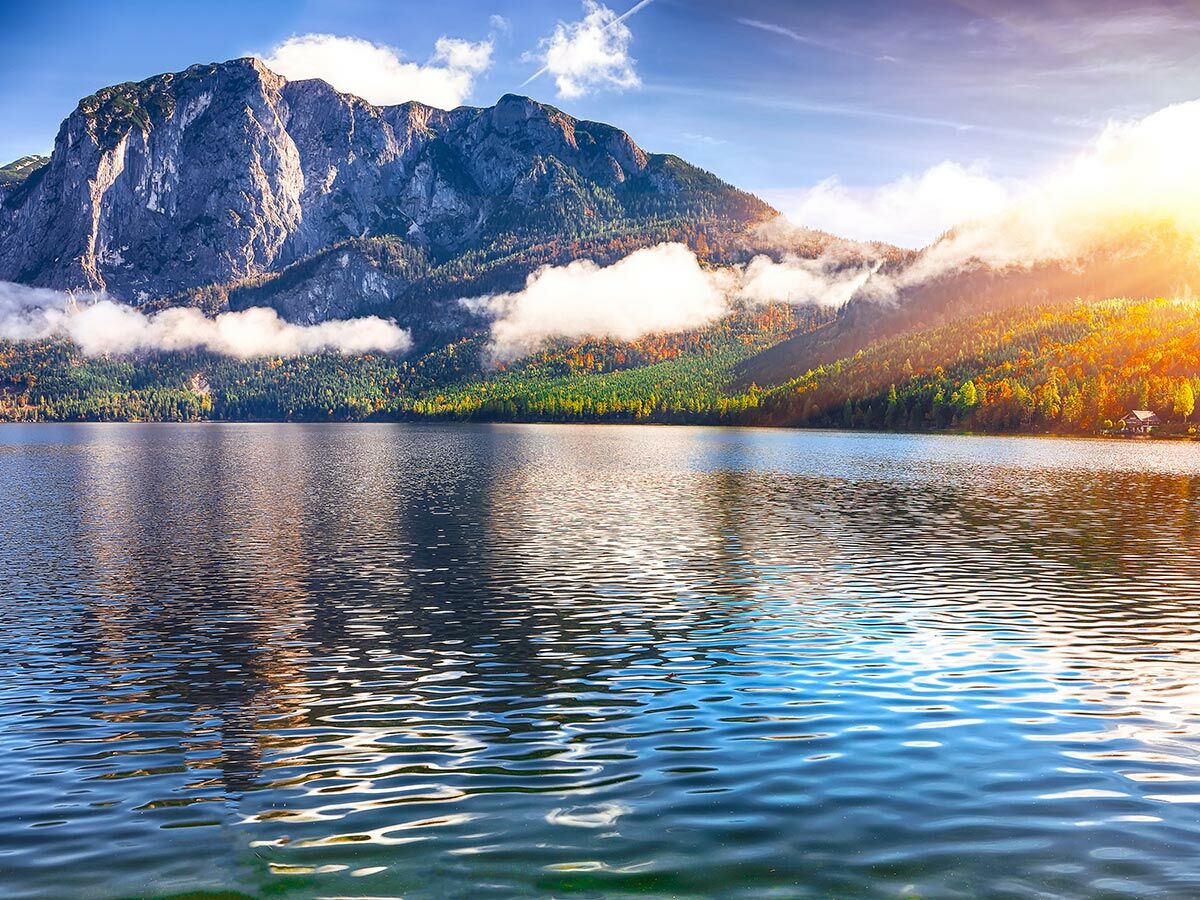 Atemberaubende Berglandschaft im Herbst mit See, Bäumen in Herbstfarben, Wolken und strahlendem Sonnenlicht am Horizont.
