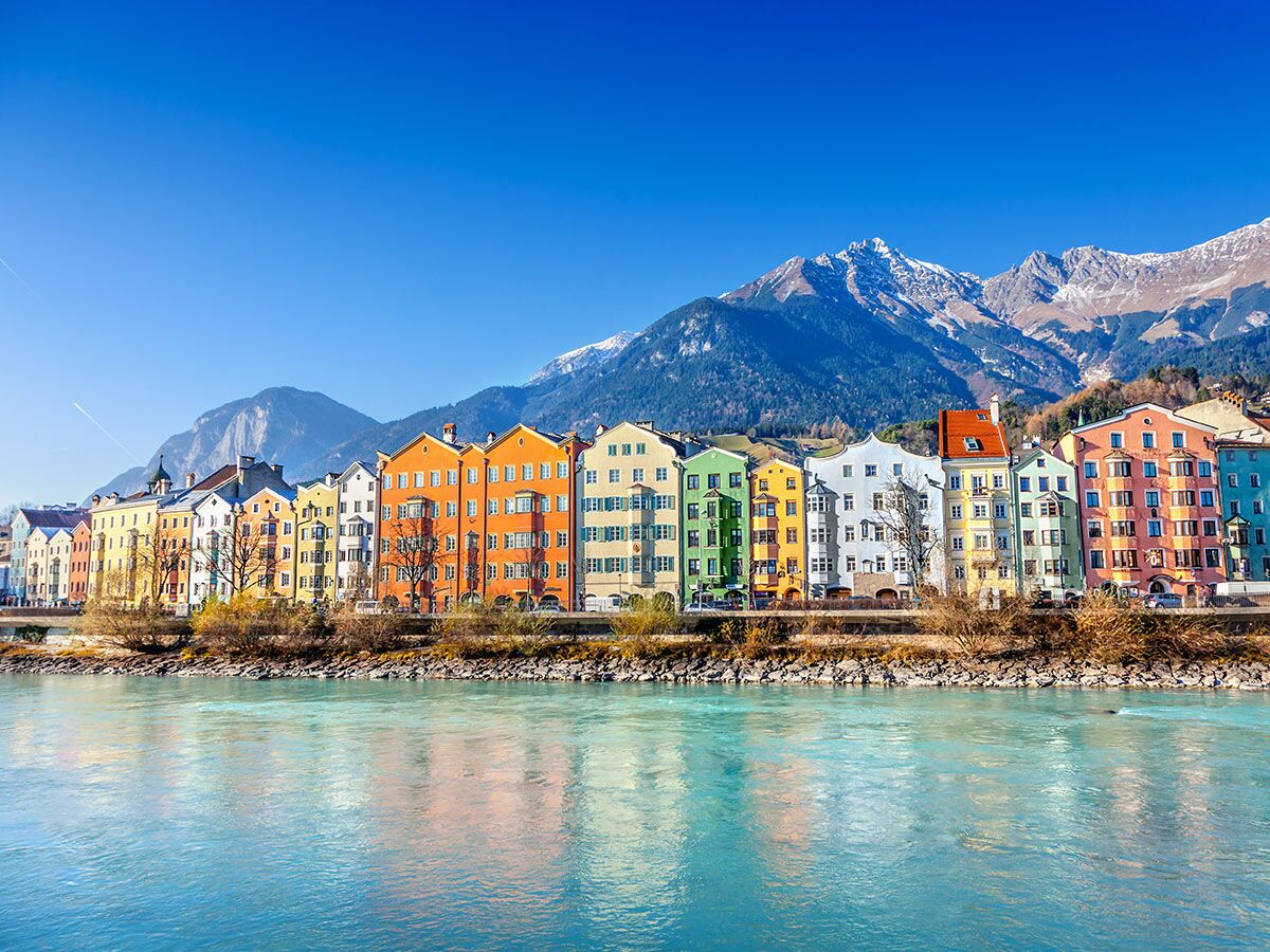 Reihe leuchtend bunter Häuser am Inn Fluss in Innsbruck, Österreich, mit schneebedeckten Alpen und blauem Himmel.