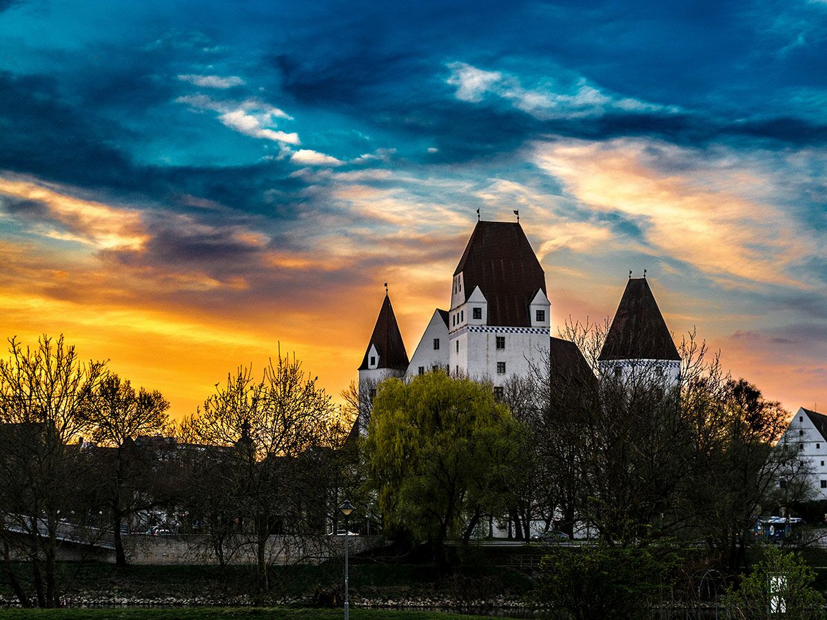 Historisches Schloss Ingolstadt bei Sonnenuntergang mit orange-blauem Himmel und Bäumen im Vordergrund.