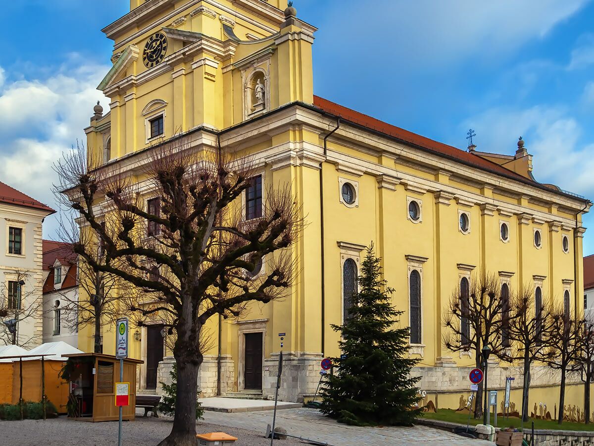 Große gelbe Barockkirche mit Uhr, umgeben von kargen Bäumen und Weihnachtsbaum auf einem winterlichen Marktplatz.