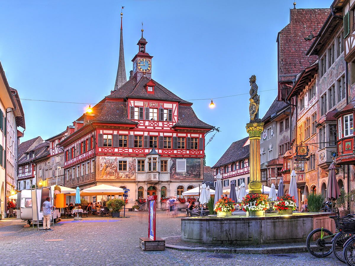 Historischer Rathausplatz in Stein am Rhein bei Abenddämmerung mit Brunnen, geschäftigen Cafés und bunt bemalten Gebäuden.