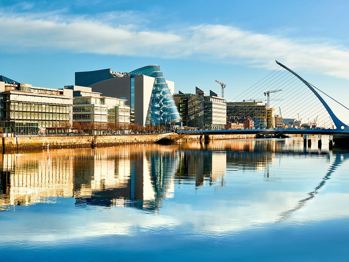 Modernes Dubliner Stadtbild am Fluss Liffey mit der Samuel Beckett Brücke und Gebäuden, die sich im Wasser spiegeln, unter blauem Himmel.
