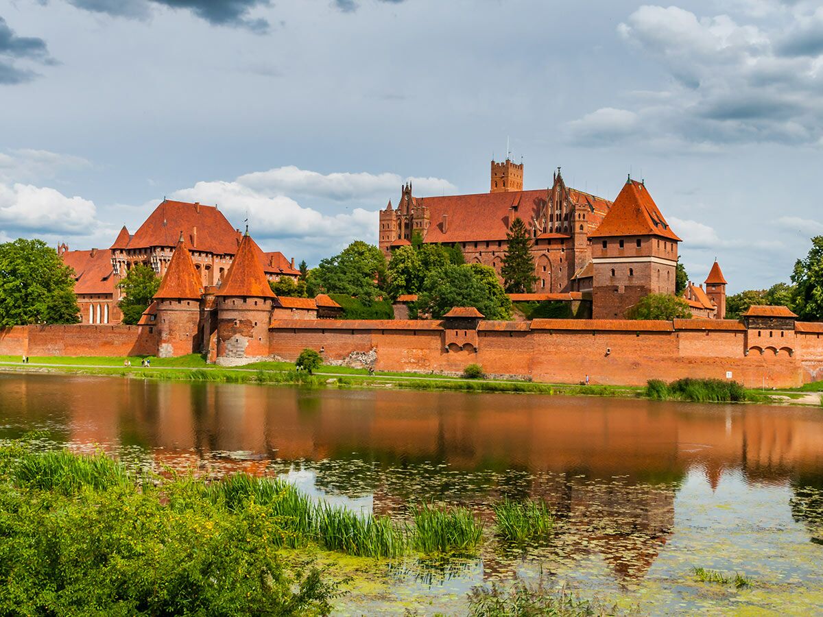 Majestätische Malbork Burg aus rotem Backstein mit Dächern und Türmen, gespiegelt im Fluss unter bewölktem Himmel.