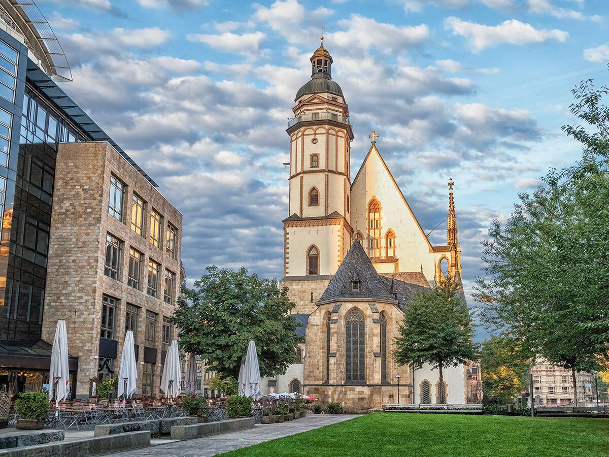 Die Thomaskirche in Leipzig neben einem modernen Gebäude mit Glasfassade, einem Außencafé und grünem Rasen bei Tageslicht.