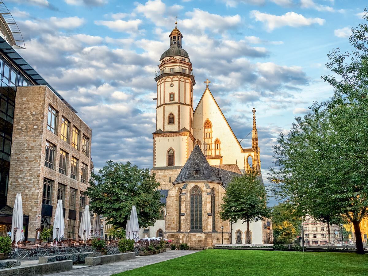 Panoramablick auf die Thomaskirche in Leipzig mit modernem Gebäude, Außenbereich und grünem Park an einem sonnigen Tag.