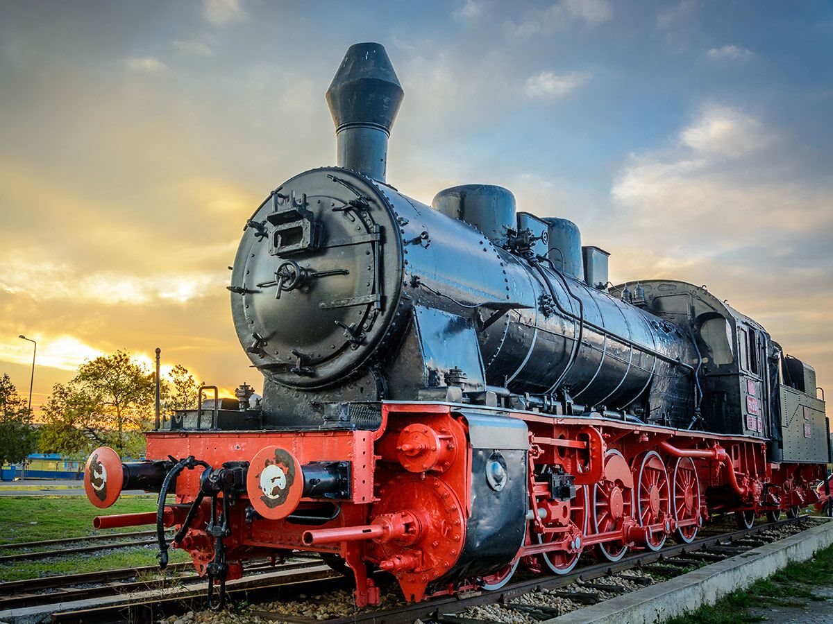 Historische schwarze Dampflokomotive mit roten Rädern und Details steht bei Sonnenuntergang auf Bahngleisen.