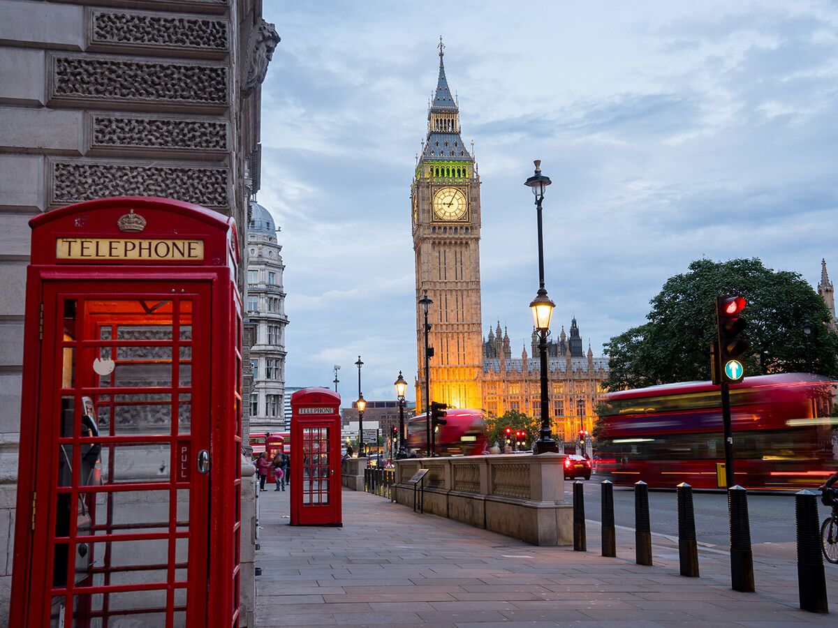 Ikonische rote Londoner Telefonzelle vor Big Ben und dem Parlament bei Dämmerung mit Lichtspuren von Bussen auf der Straße.