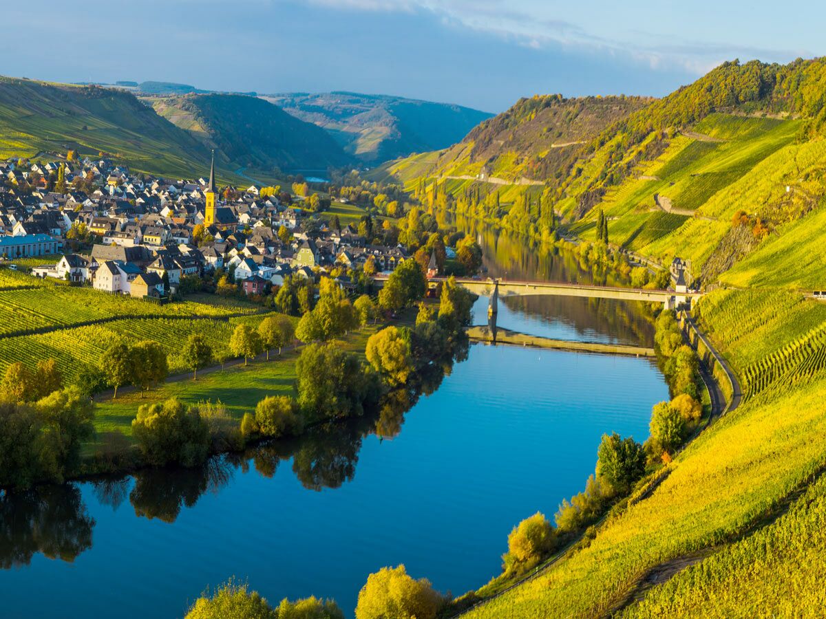 Malerisches Dorf im Moseltal mit Fluss, Brücke und gelben Weinbergen unter blauem Himmel im Herbst.
