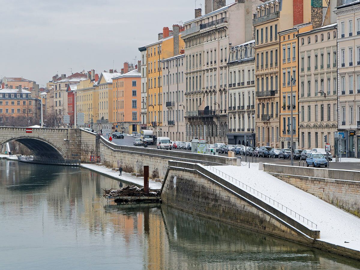 Winterliche Stadtansicht von Lyon mit Fluss, Steinbrücke, bunten Gebäuden, Autos und Schnee am Ufer.