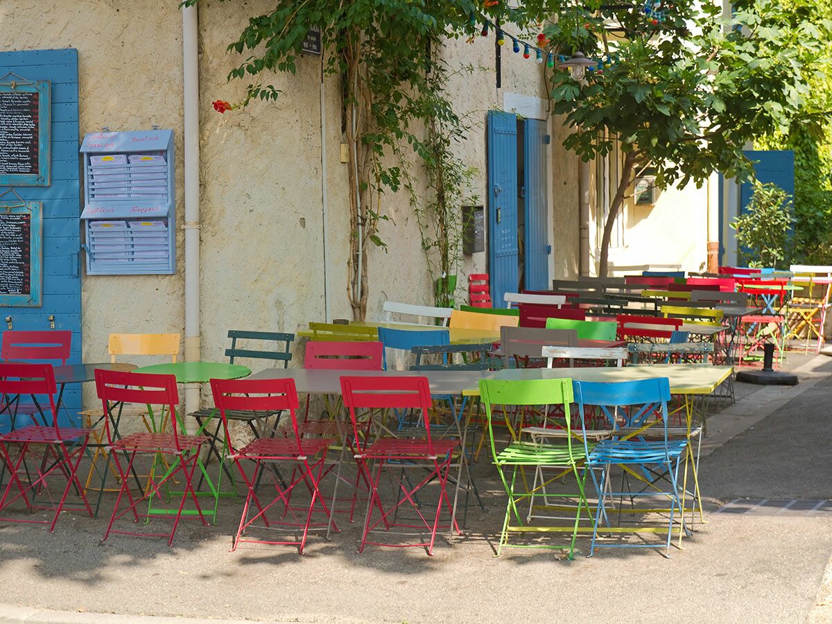 Reihe von leeren, bunten Metallstühlen und Tischen auf einer sonnigen Straßenterrasse eines mediterranen Cafés.