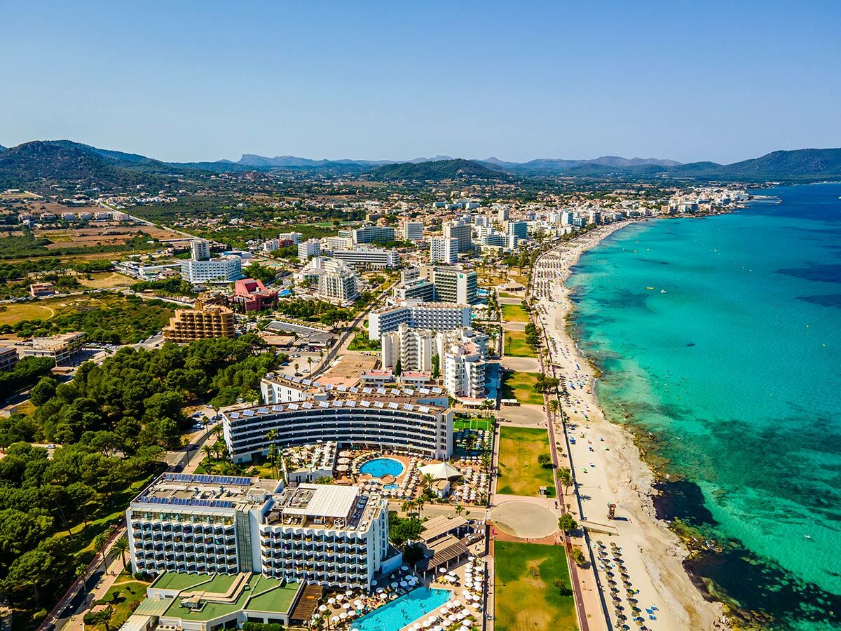 Luftaufnahme einer sonnigen Küstenstadt auf Mallorca mit langem Sandstrand, türkisfarbenem Meer und vielen Hotels.