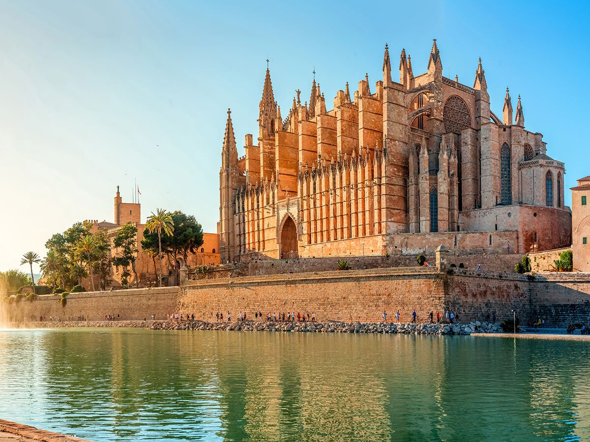 Die imposante Kathedrale von Palma de Mallorca am Wasser, mit Menschen am Ufer, im warmen Licht der goldenen Stunde.