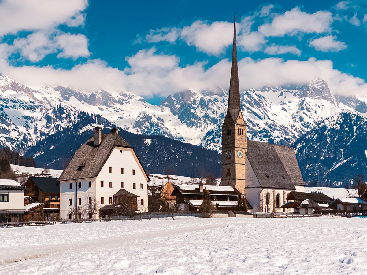 Verschneites Alpendorf mit spitzer Kirche, traditionellem Haus und schneebedeckten Alpen im Hintergrund bei blauem Himmel.