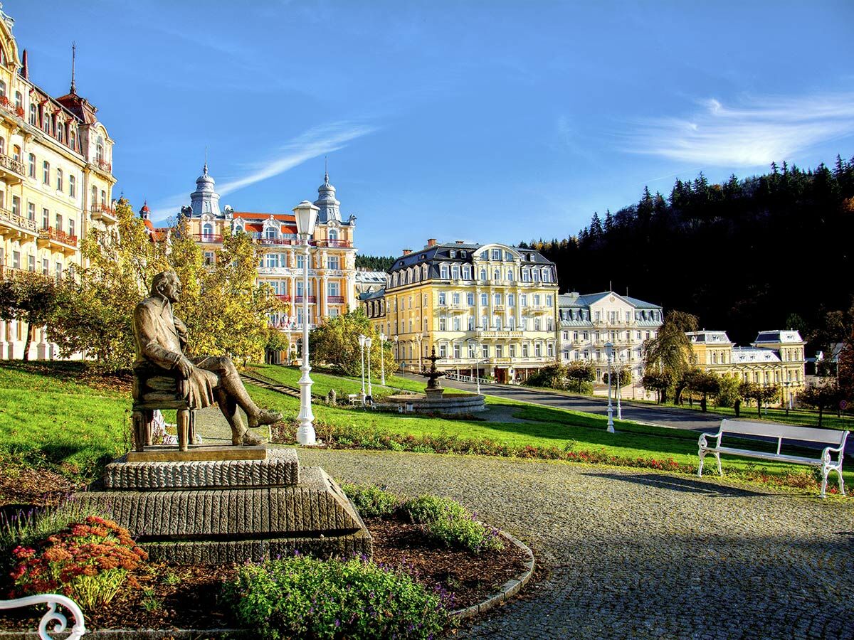 Bronze-Statue im historischen Kurpark umgeben von eleganten Hotels, Bäumen und Wegen an sonnigem Herbsttag.