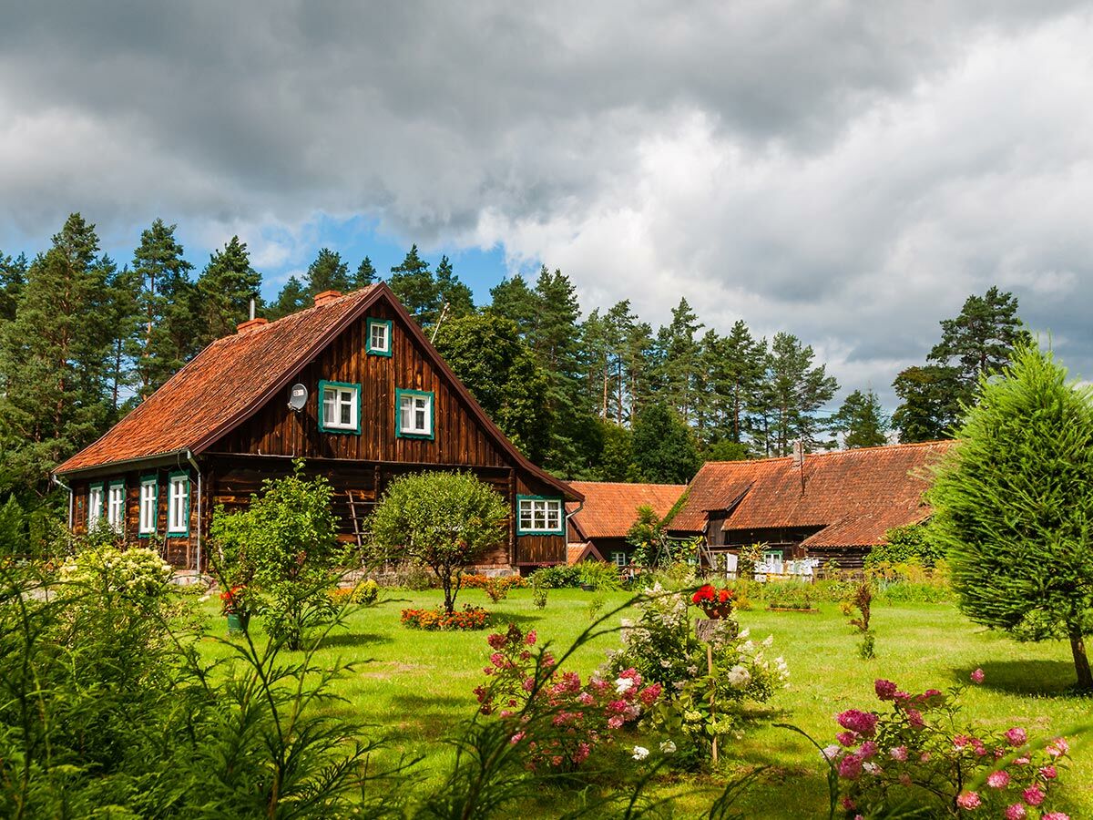 Traditionelles Holzhaus mit rotem Dach, grünen Fenstern und bunten Blumen im Garten unter bewölktem Himmel.