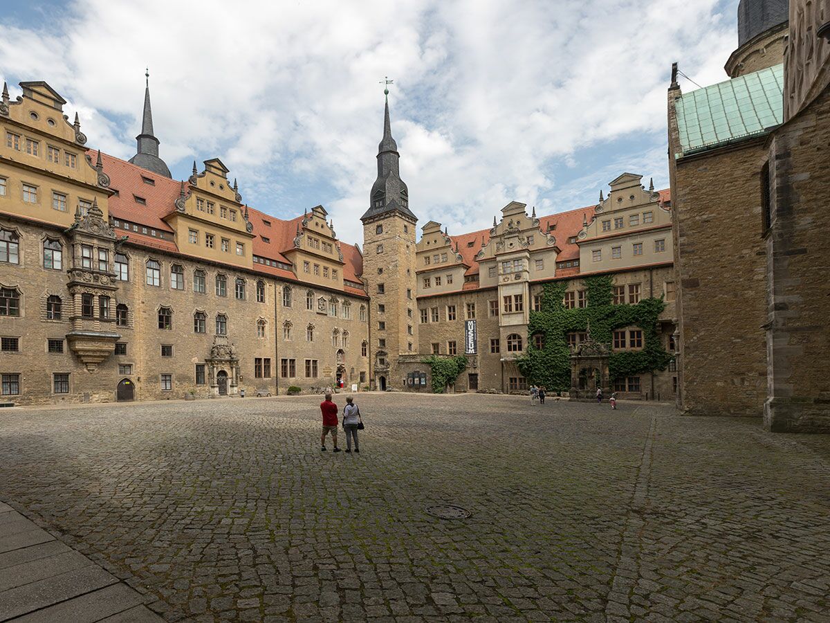 Historischer Innenhof von Schloss Altenburg mit Sandsteingebäuden, roten Dächern, spitzen Türmen, Efeu und Museumsschild. Zwei Personen im Vordergrund.