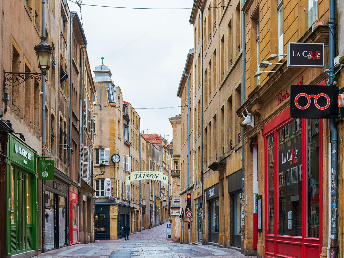 Historische Rue Taison in Metz, Frankreich, mit alten Gebäuden, Geschäften wie 'Palais des Thés' und 'La Cave' unter bewölktem Himmel.