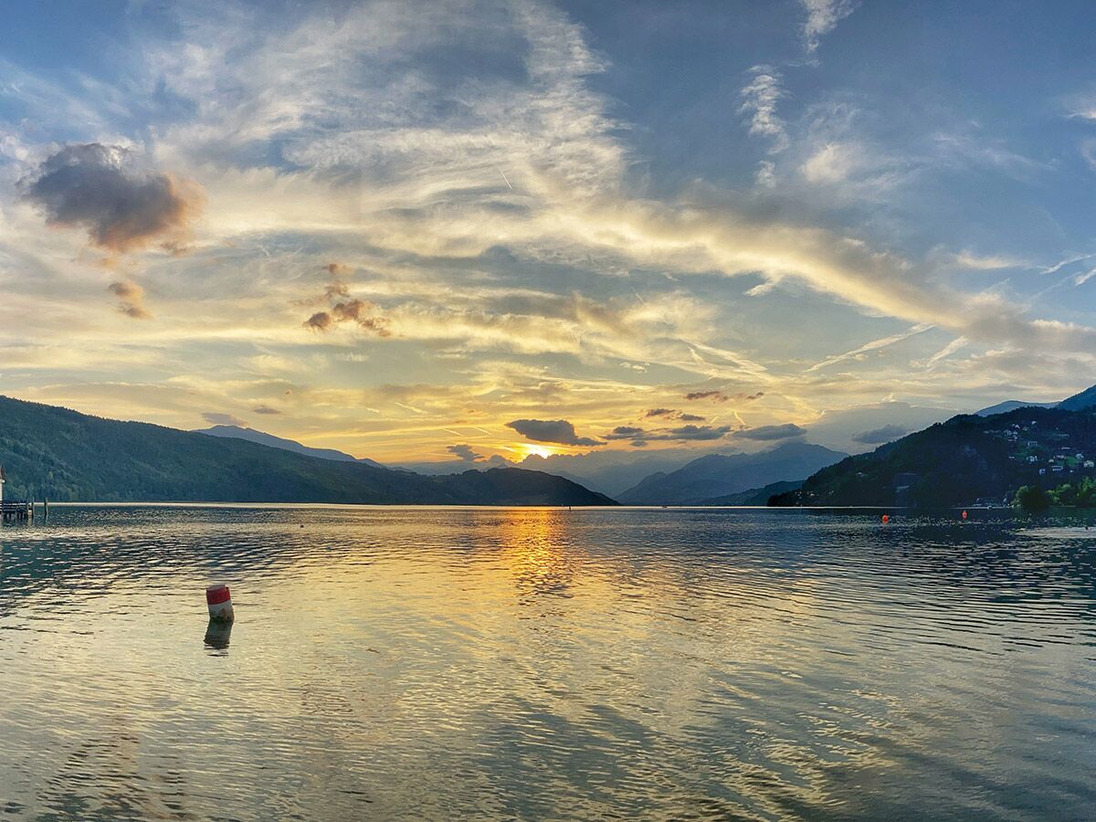 Malerischer Sonnenuntergang über einem ruhigen See mit goldenen Reflexionen, umgeben von bewaldeten Bergen unter einem bewölkten Himmel.