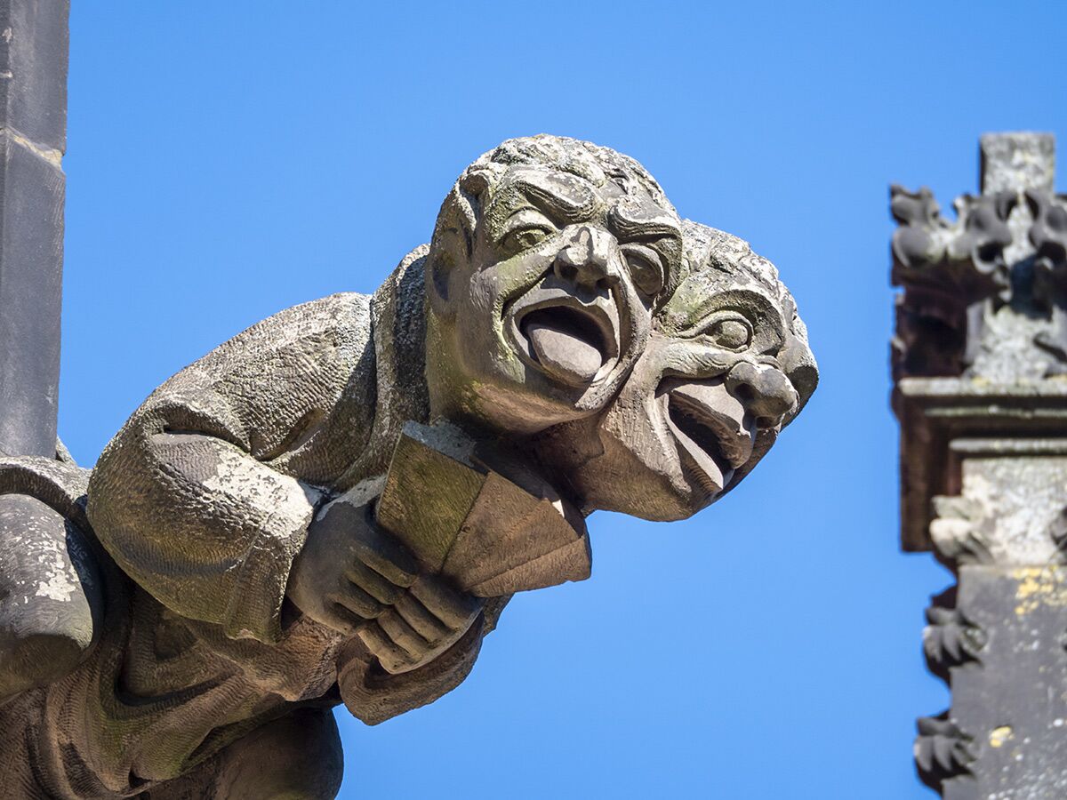 Detailaufnahme eines gotischen Wasserspeiers mit zwei expressiven Gesichtern aus Stein vor blauem Himmel.