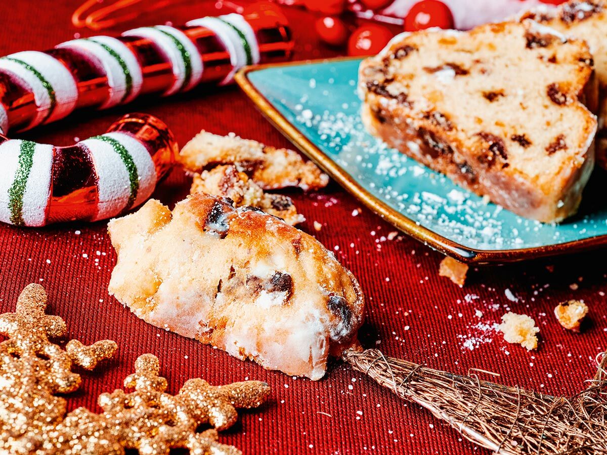 Scheiben von Stollen und Weihnachtskuchen mit Puderzucker auf rotem Grund, umgeben von festlicher Deko wie Zuckerstangen und Schneeflocken.