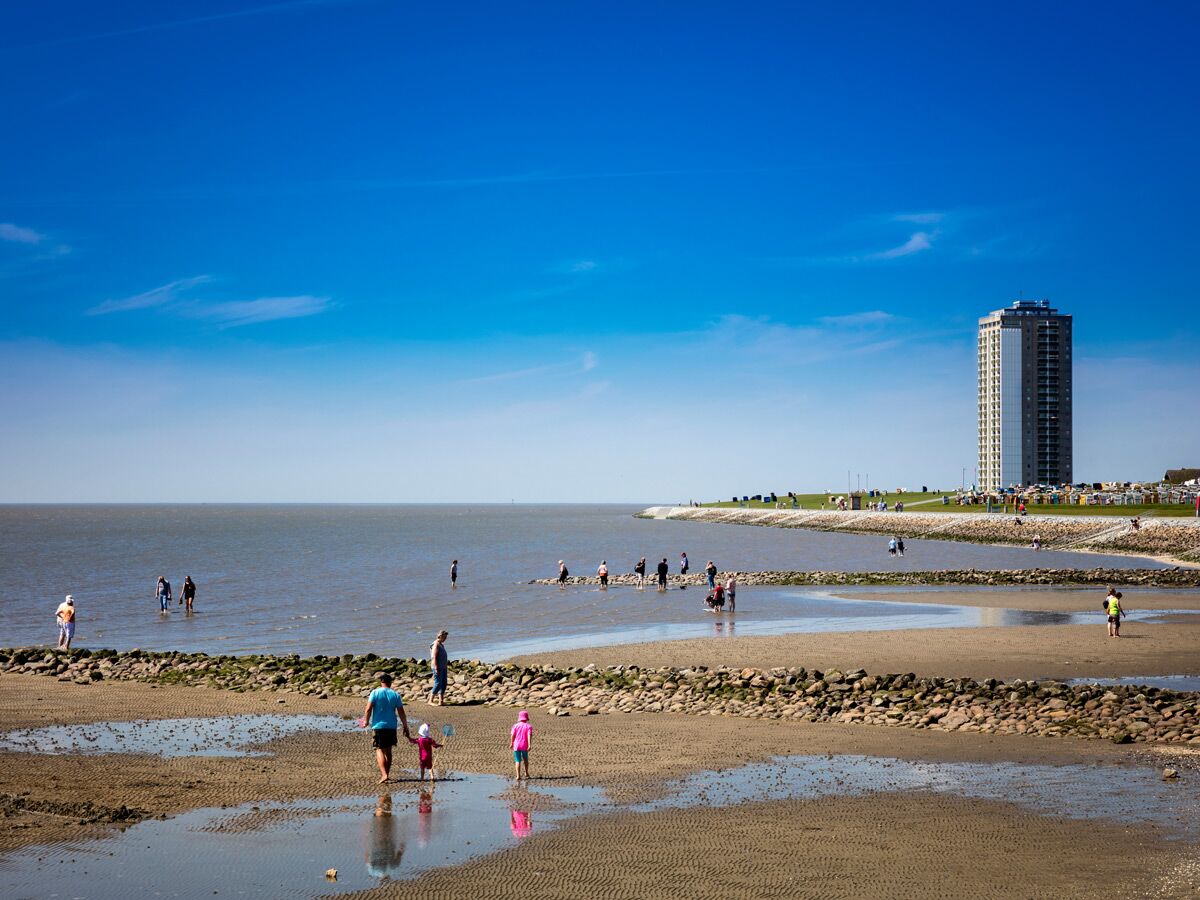 Familien und Kinder erkunden den Strand bei Ebbe an der Nordseeküste, mit modernem Hochhaus und Strandkörben im Hintergrund.