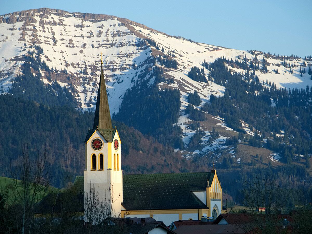 Majestätische Alpenkirche mit hohem Kirchturm und roten Uhren, davor schneebedeckte Berge und grüne Wälder im Sonnenlicht.