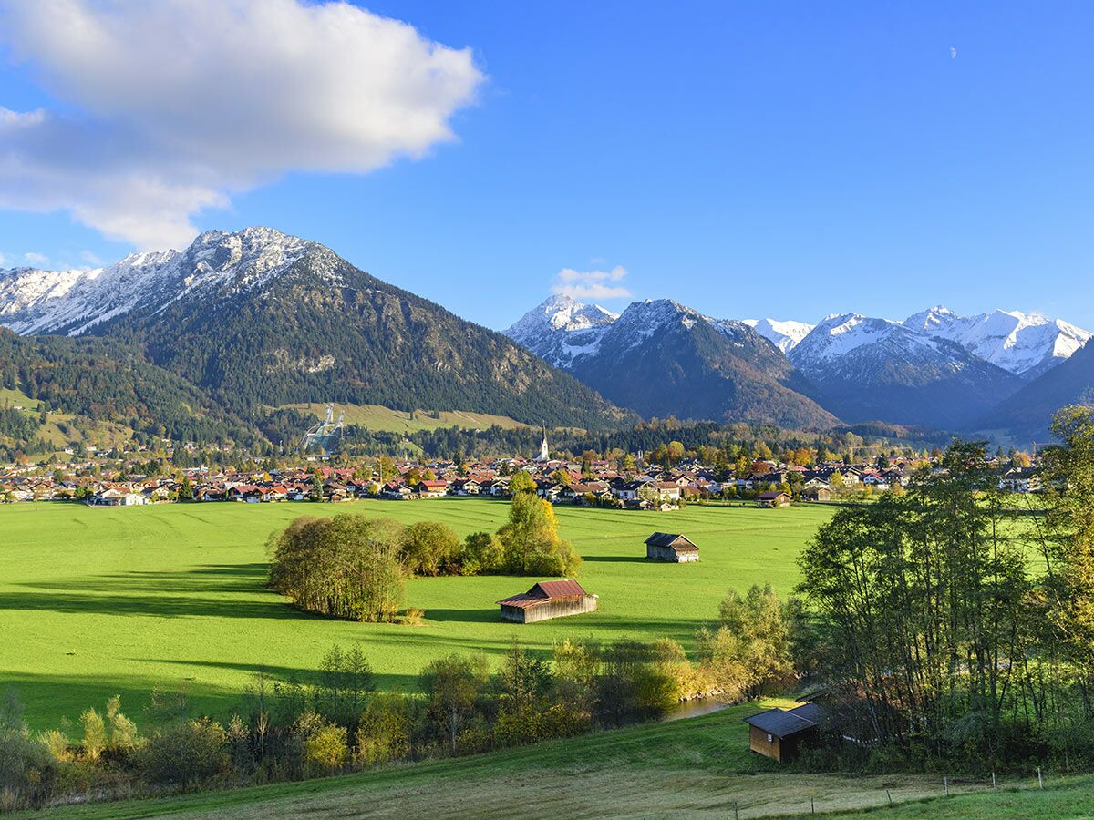 Malerisches Bergdorf in den Alpen, umgeben von grünen Wiesen und schneebedeckten Gipfeln unter blauem Herbsthimmel.