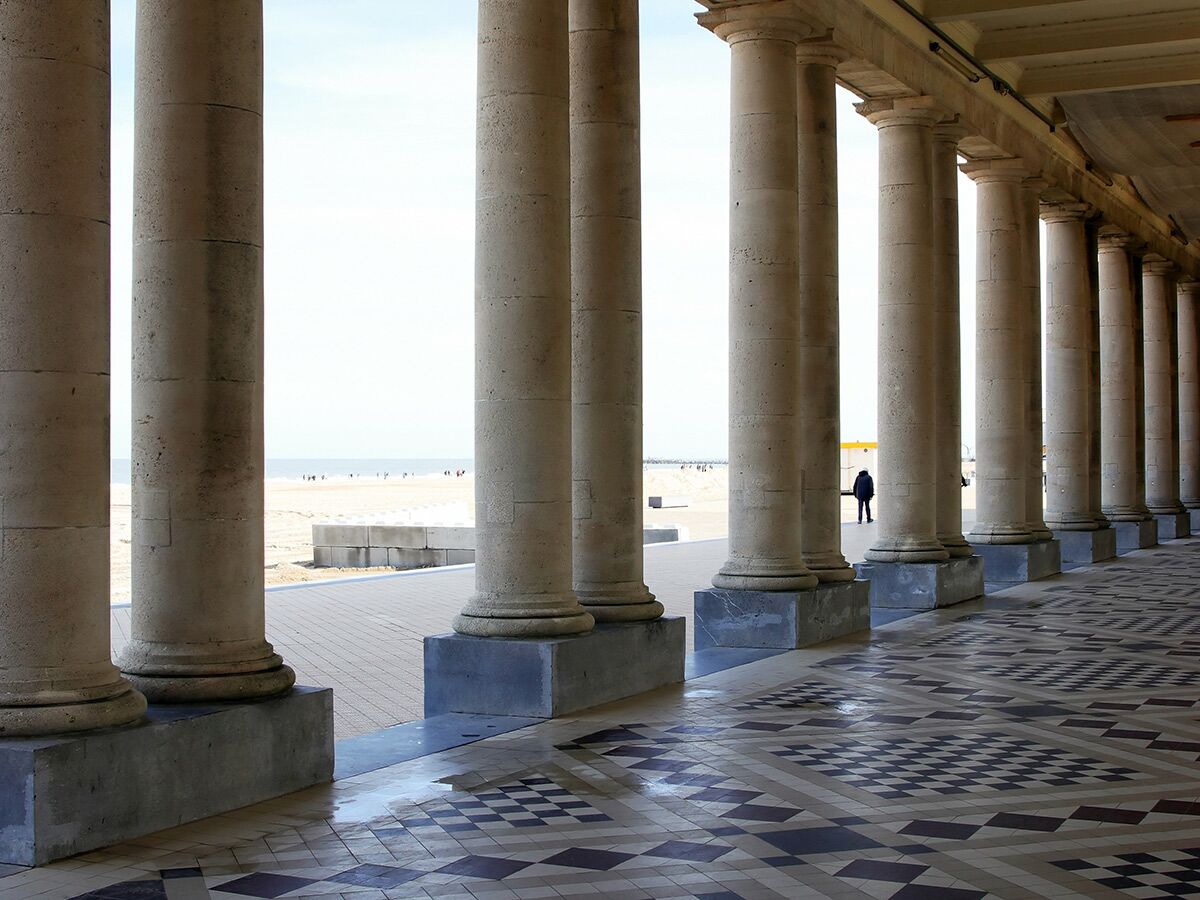 Lange Promenade mit steinernen Säulen bietet Blick auf sonnigen Strand und Meer. Eine Person steht am Ende des Ganges.