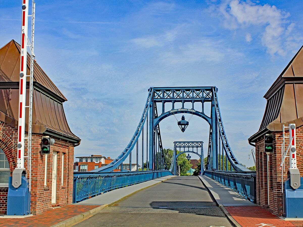 Historische Kaiser-Wilhelm-Brücke in Wilhelmshaven, Deutschland, mit blauen Stahlbögen, Backsteinhäusern und Schranken bei Tag.