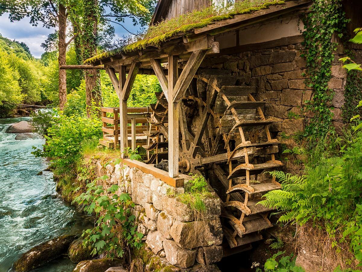 Historische Wassermühle mit großem Holzrad am rauschenden Fluss, umgeben von üppigem Grün und Wald.
