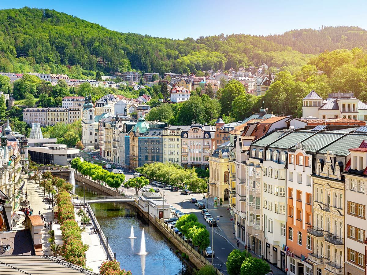 Panoramablick auf Karlovy Vary mit dem Teplá-Fluss, bunten historischen Gebäuden, grünen Hügeln und blauem Himmel.