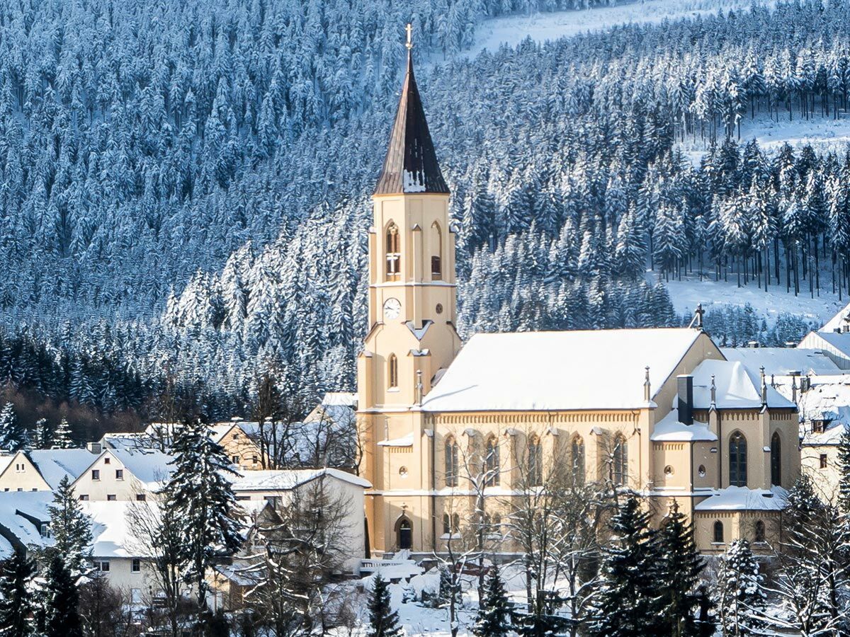 Majestätische Kirche mit hohem Turm und schneebedecktem Dach, eingebettet in ein winterliches Dorf und dichten, verschneiten Wald.
