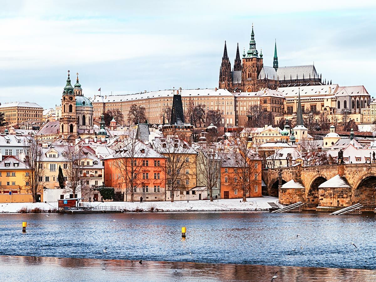 Prag im Winter: verschneite Dächer, Karlsbrücke, Moldau und Prager Burg mit Veitsdom unter bewölktem Himmel.