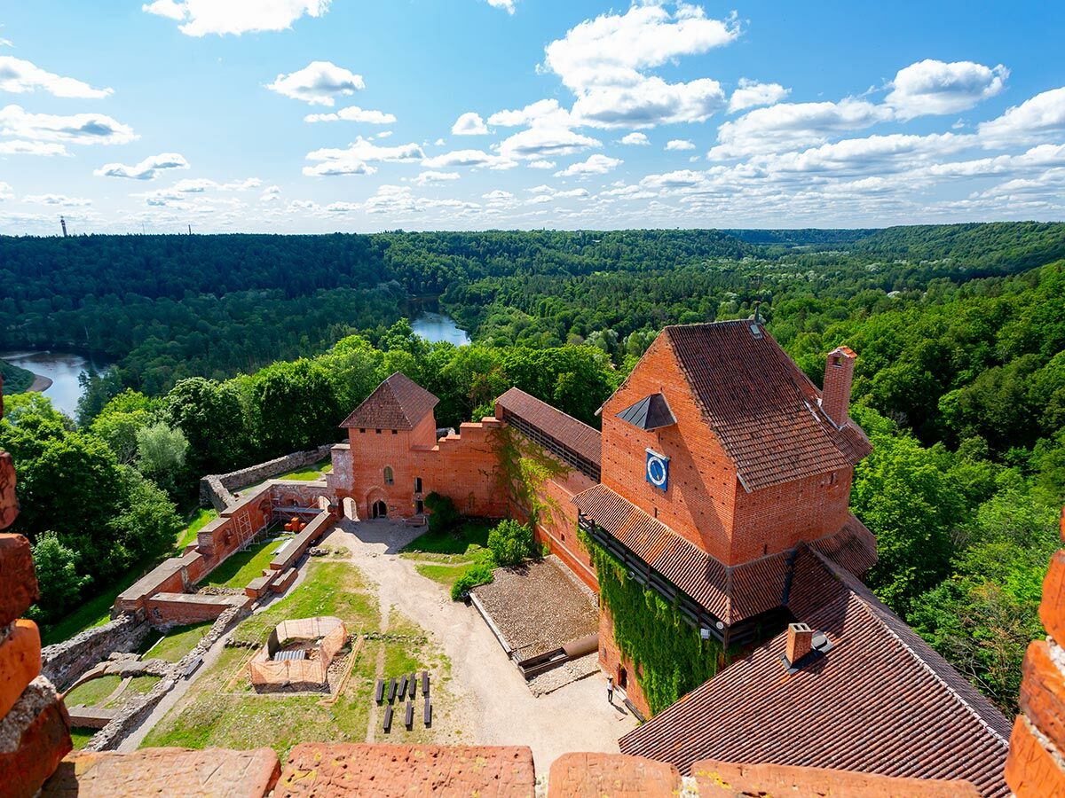 Rote Backsteinburg Turaida in Lettland, umgeben von einem grünen Wald und dem gewundenen Fluss Gauja unter blauem Himmel.