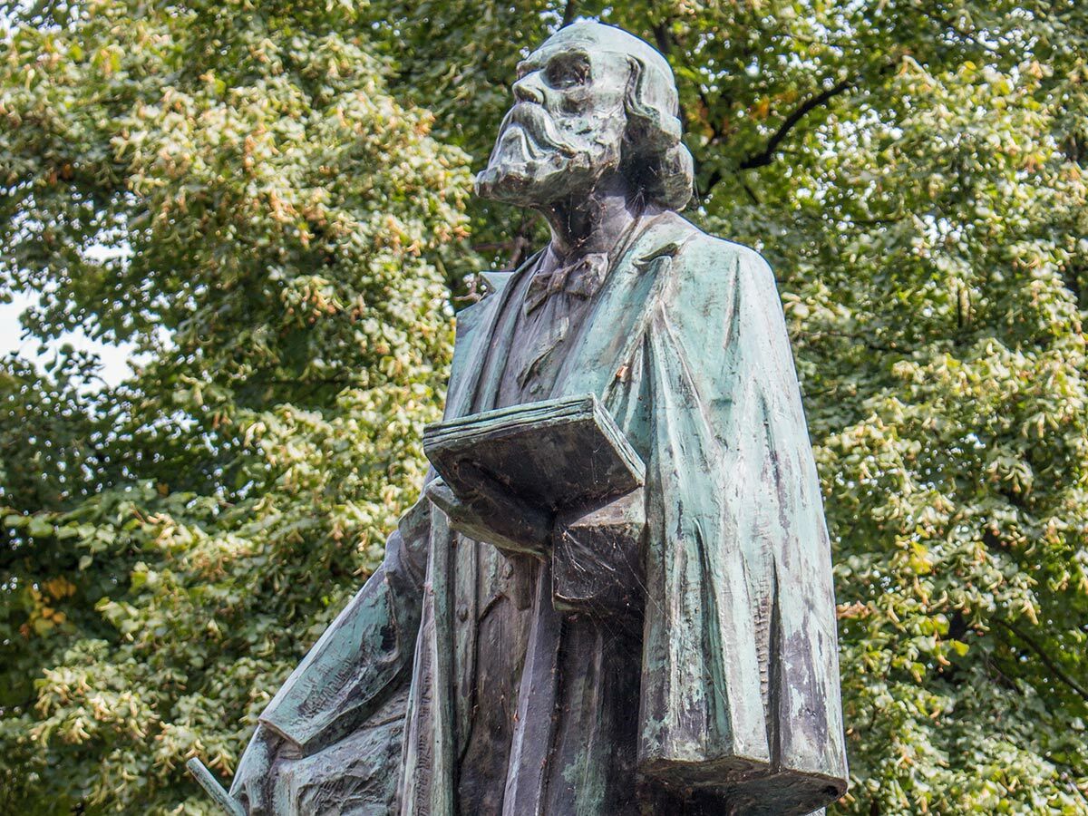 Bronzestatue eines bärtigen Mannes mit Buch, blickt auf. Grüne Patina, im Freien vor Bäumen.