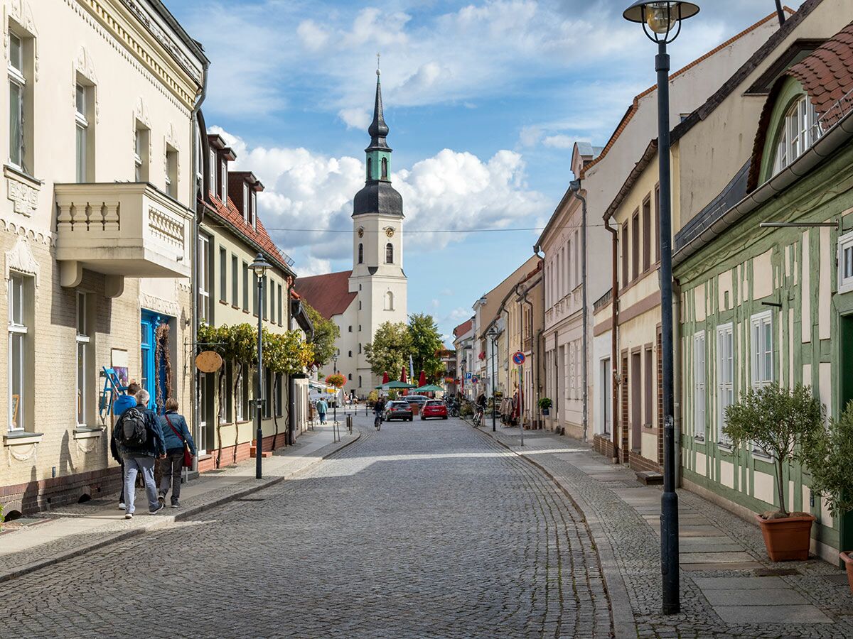 Kopfsteinpflasterstraße in historischer Altstadt mit Fachwerkhäusern, Fußgängern und Kirchturm unter blauem Himmel.