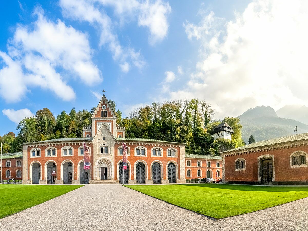 Historisches Hofmarstall Gebäude in Salzburg mit roter Ziegelfassade, Bogenfenstern, Uhrturm, umgeben von grünen Wiesen und Bergen.