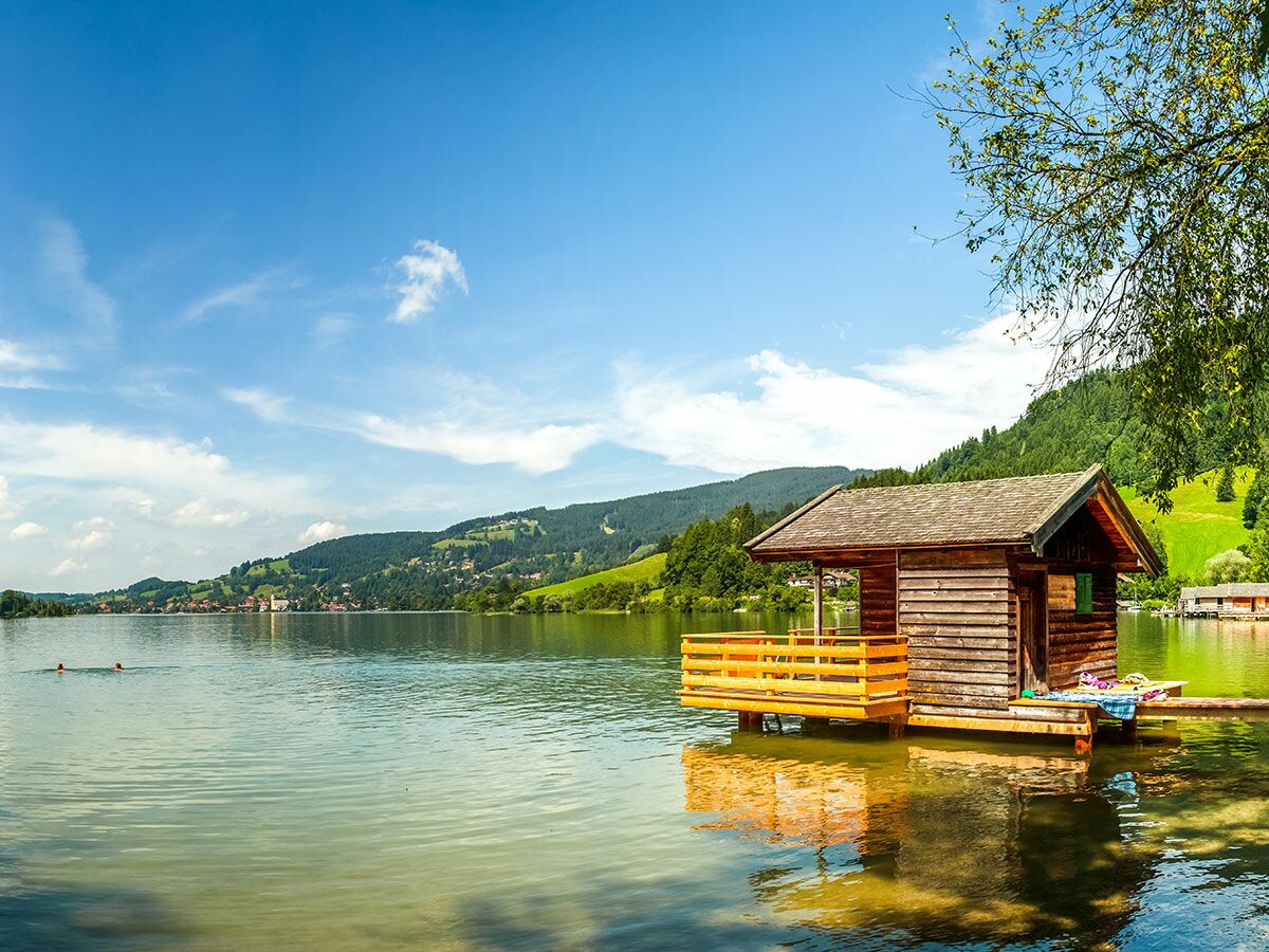 Idyllisches Holzhaus mit Steg auf einem Bergsee, umgeben von grünen Hügeln und Bergen, bei Sonnenschein. Zwei Schwimmer im Wasser.