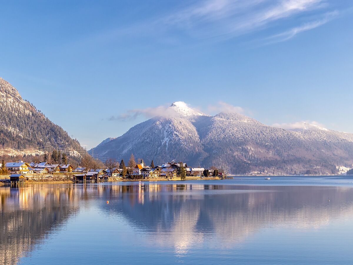 Idyllisches Alpendorf am Seeufer mit schneebedeckten Bergen und klarem blauen Himmel im Winter, Spiegelung im ruhigen Wasser.