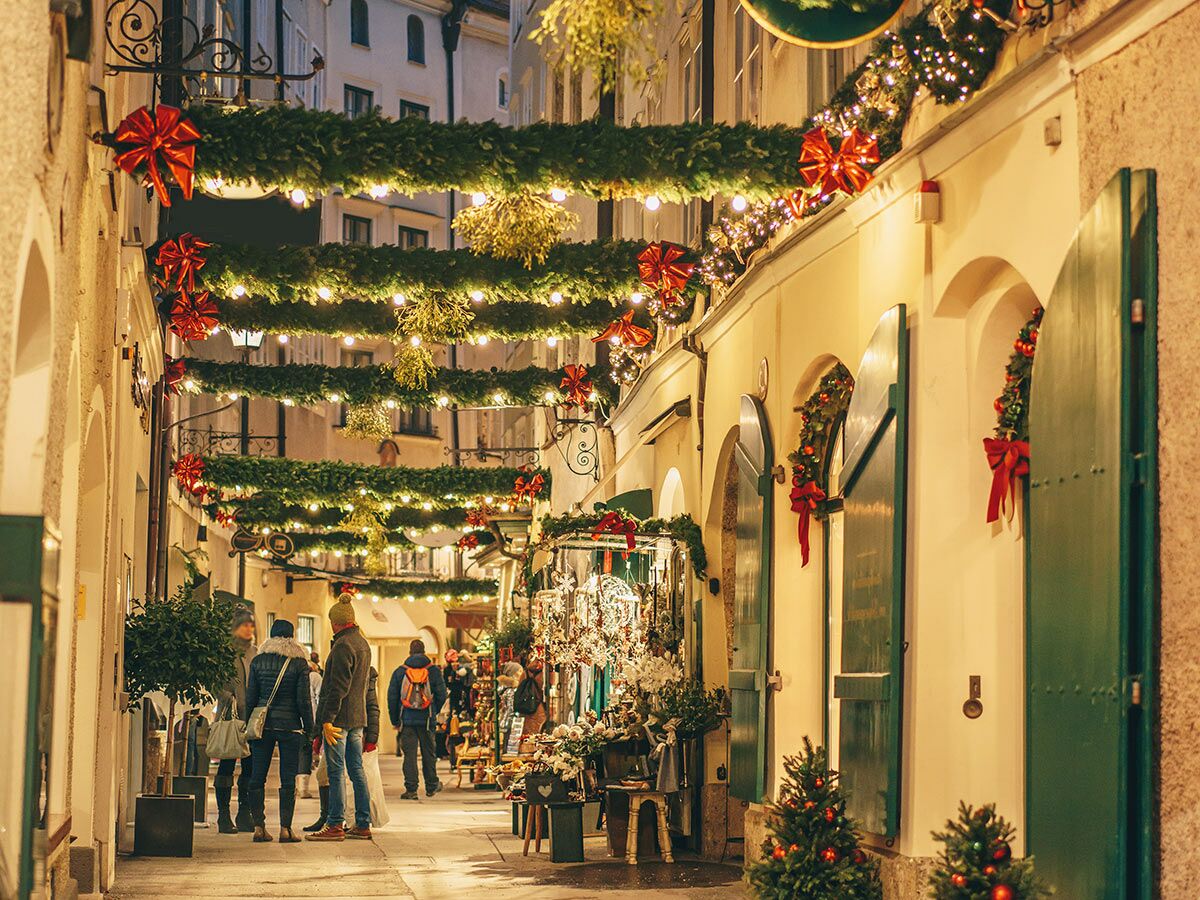 Festlich beleuchtete Weihnachtsgasse mit Lichterketten, roten Schleifen und grünen Girlanden. Passanten schlendern zwischen dekorierten Geschäften.