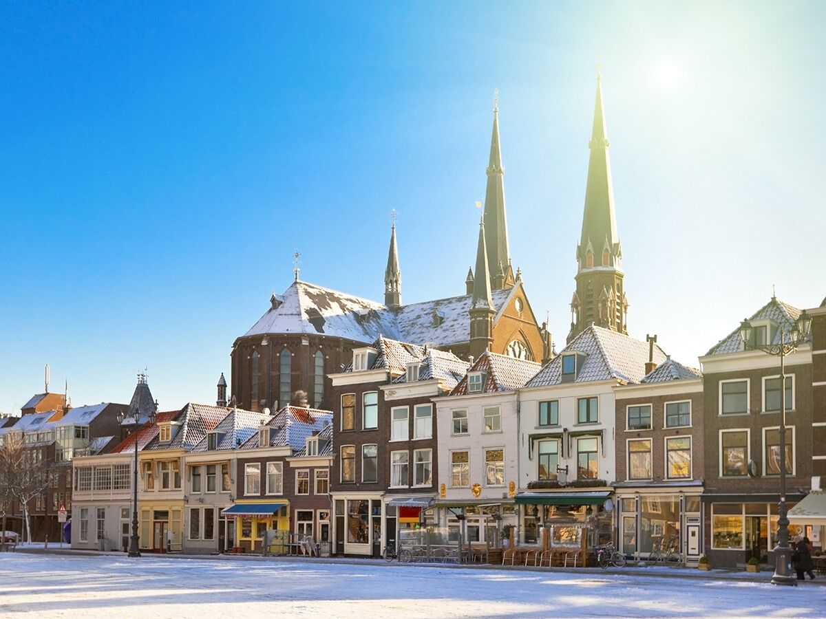 Verschneite Altstadtgasse mit historischen Gebäuden, Geschäften und zwei hohen Kirchen unter blauem Winterhimmel in Delft, Niederlande.