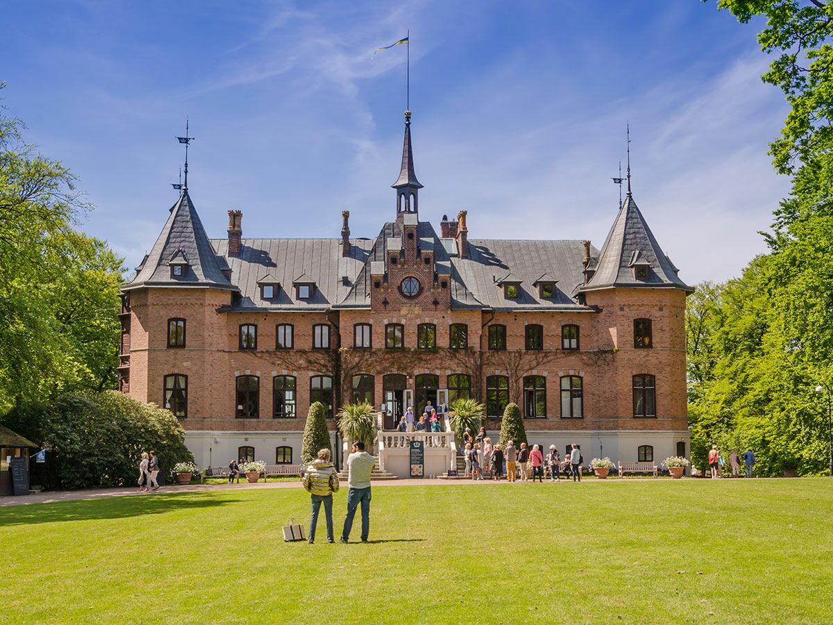 Rotes Backsteinschloss mit Türmen und Spitzdach, umgeben von grünem Park und Bäumen unter blauem Himmel mit Besuchern.